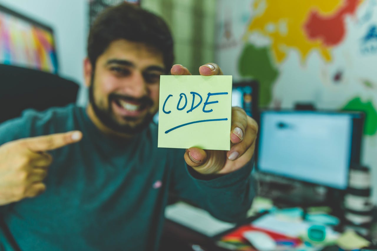 gallery-1 A smiling programmer pointing to a sticky note with 'code' written on it in an office setting.